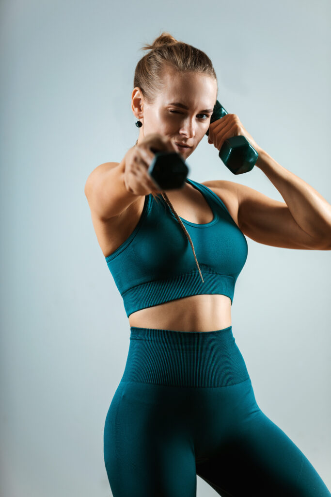 Beautiful fitness model doing exercises with dumbbells in hands on a gray background. Close-up. Woman posing on camera with dumbbells. Healthy lifestyle. Achieving goals, sports motivation. Copy space