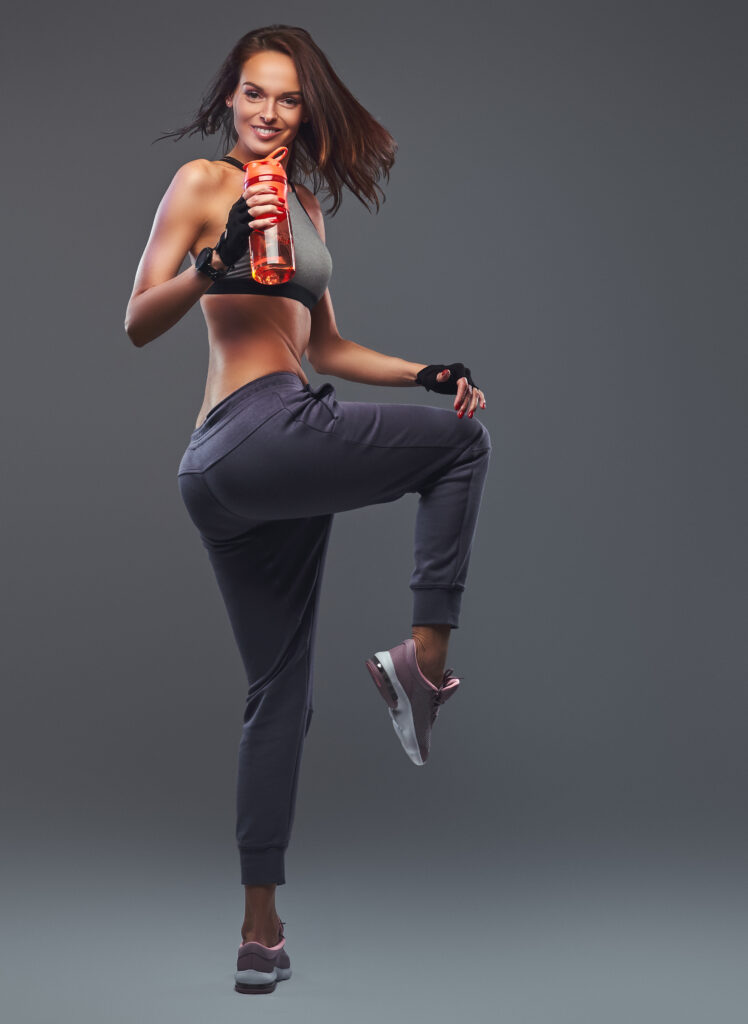Fitness brunette girl in a gray sportswear posing with a bottle in a studio.